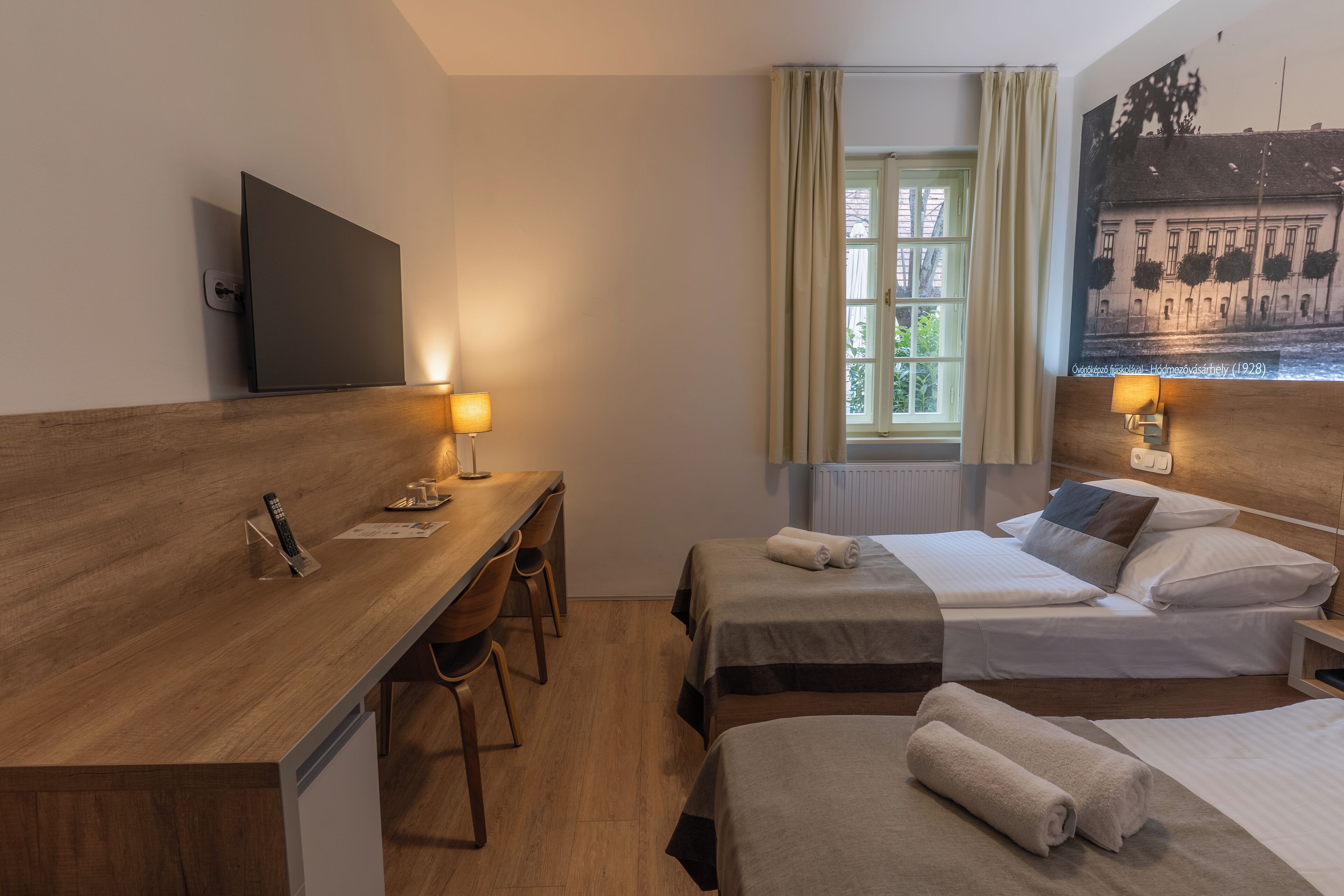 Ein Hotelzimmer mit einem Holztisch, zwei Einzelbetten, einem an der Wand montierten Fernseher und einem Fenster mit Vorhängen.