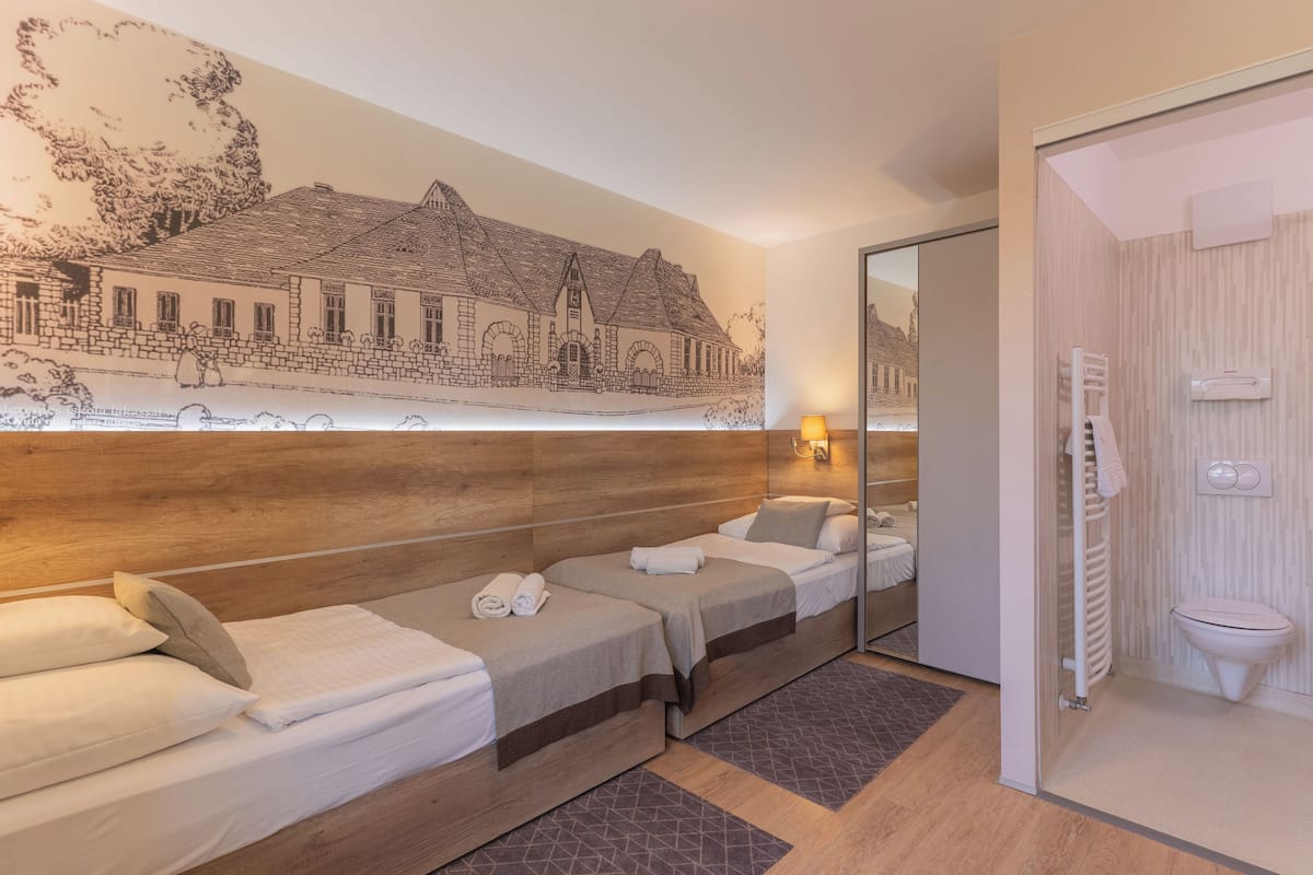 A hotel room with two single beds, a wooden headboard, a large wall mural, a mirror, a bathroom with a toilet and a towel rack.