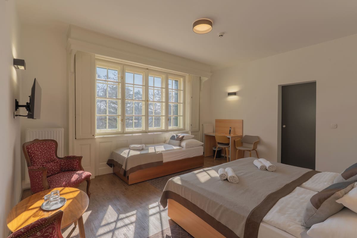 A hotel room with two beds, a desk, a chair, a red patterned armchair, and a window with a view of trees.