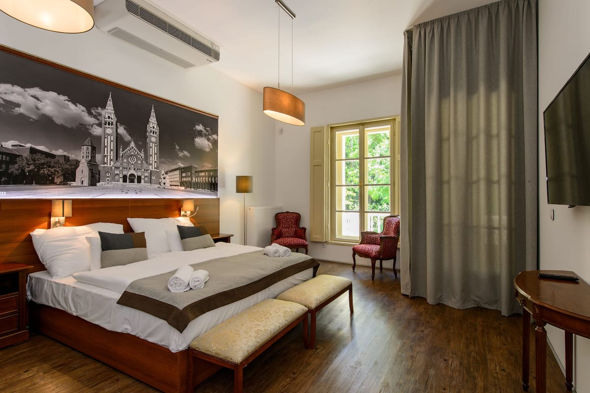 A hotel room with a large bed, a bench, a chair, a TV, and a window with curtains.