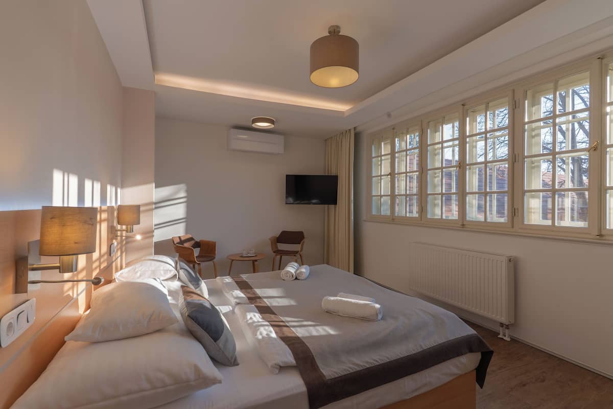 A well-lit hotel room with a large bed, a TV, a desk, and a window with a view of buildings.