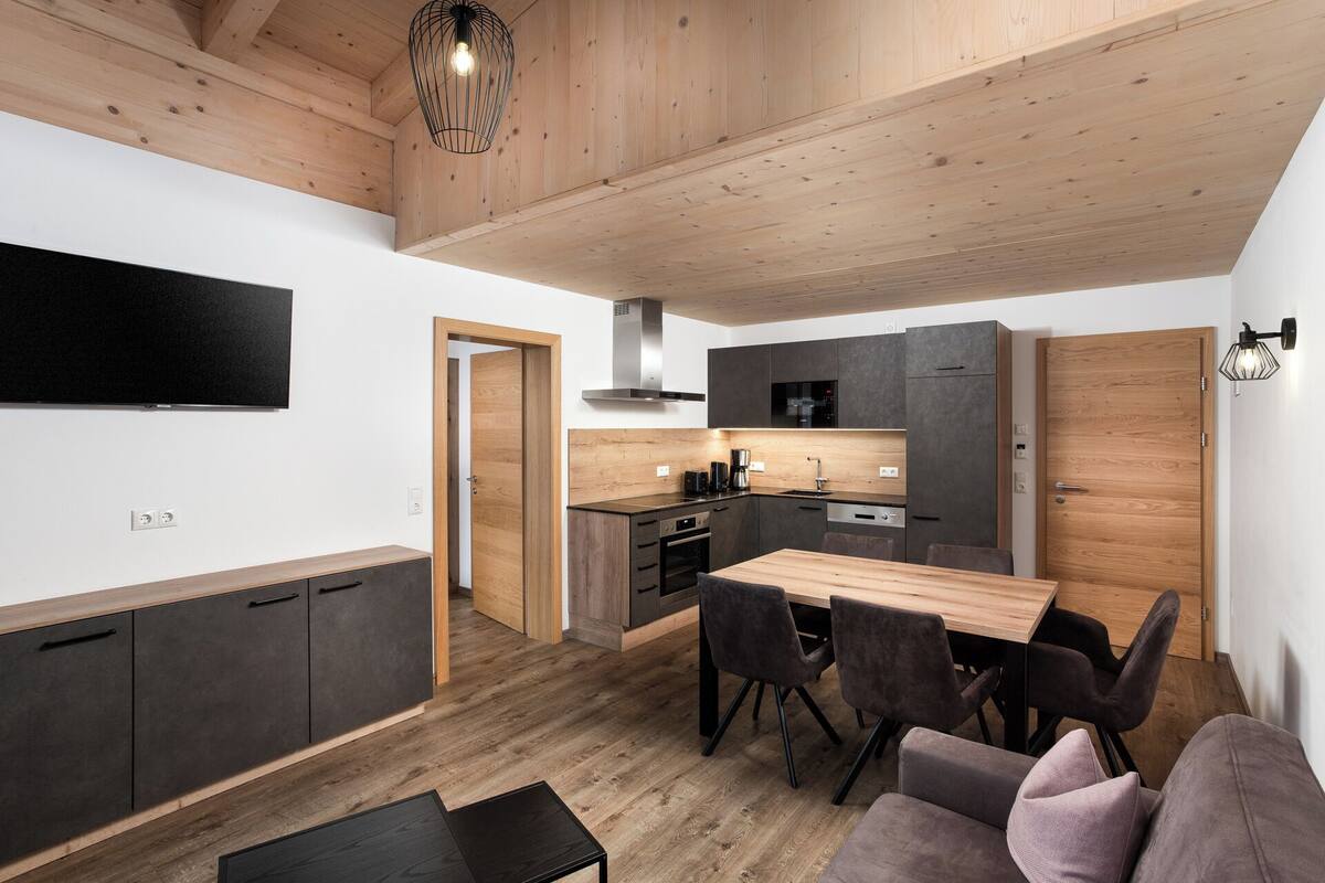 Una cocina moderna con armarios de madera, un comedor con mesa y sillas de madera, y un televisor montado en la pared.