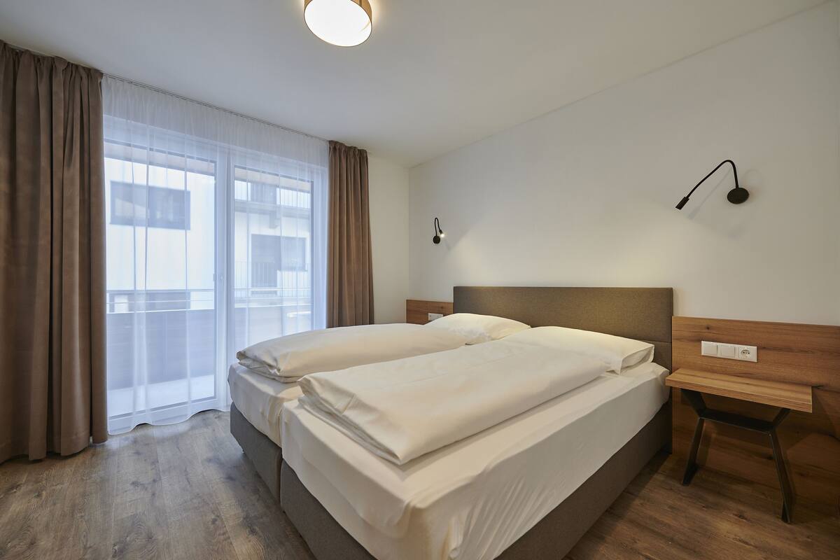 Habitación de hotel con dos camas, una mesita de noche de madera, una lámpara de pared y un ventanal con cortinas translúcidas.