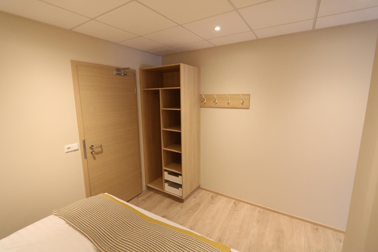 A bedroom with a wooden wardrobe, a bed with a striped blanket, and a wall-mounted light fixture.