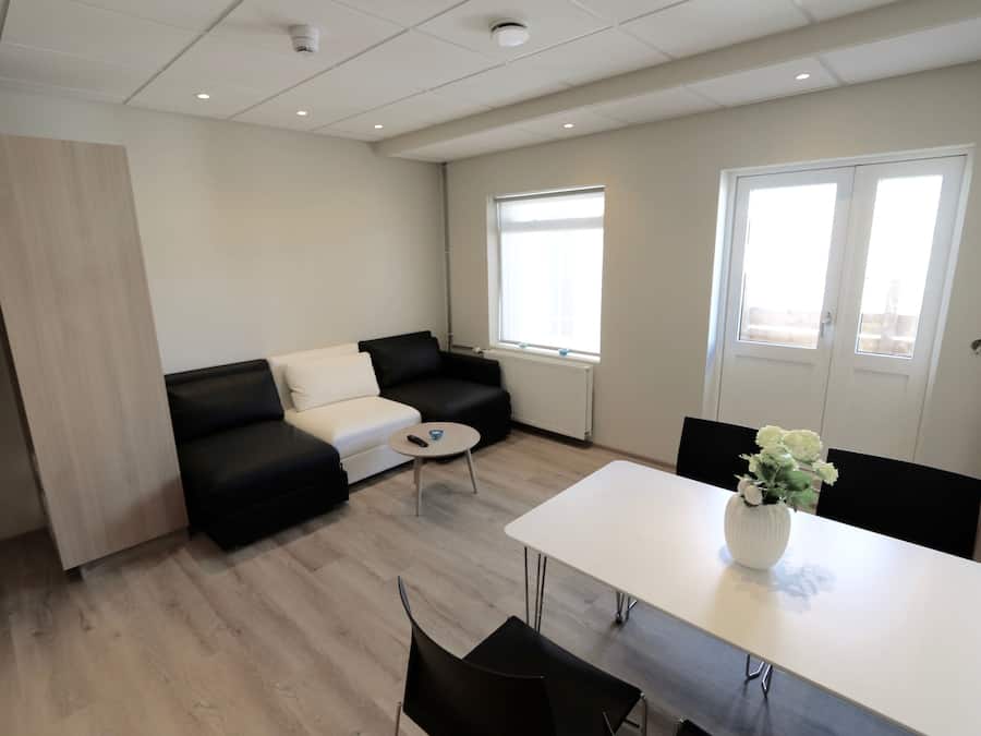 A modern living room with a black sofa, a white coffee table, and a dining table with chairs. There is a window providing natural light and a door to the right.