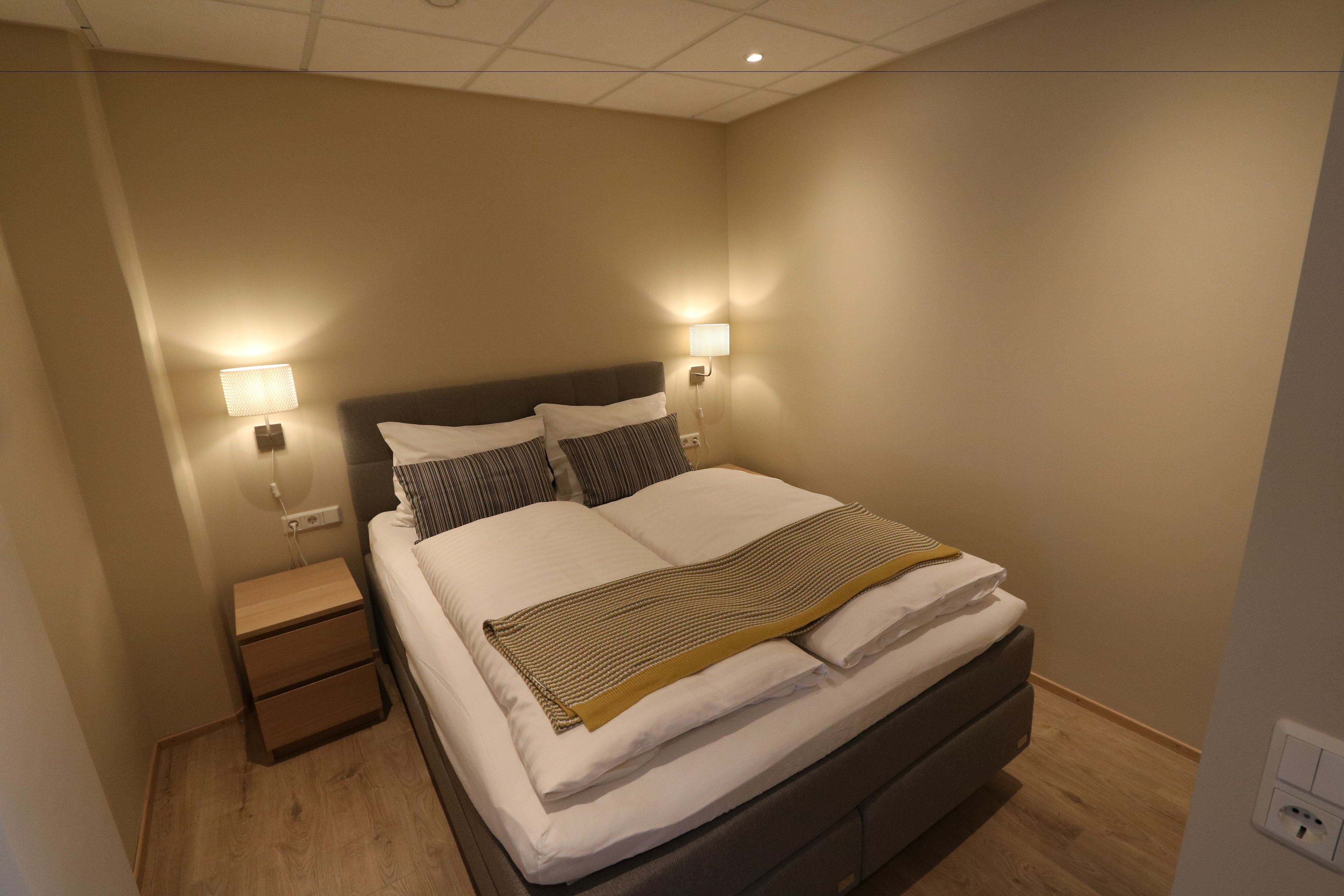 A neatly made bed with white linens and a striped blanket, two bedside lamps, a wooden nightstand, and a wall socket.