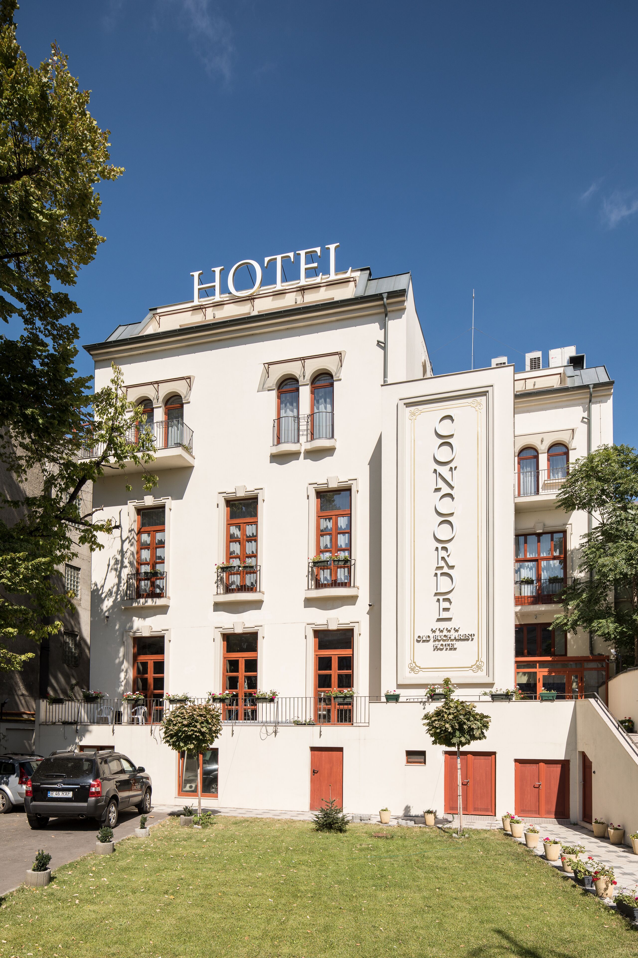 A hotel with a prominent "HOTEL" sign on the roof, a classic facade with multiple windows and balconies, and a well-maintained lawn with potted plants.