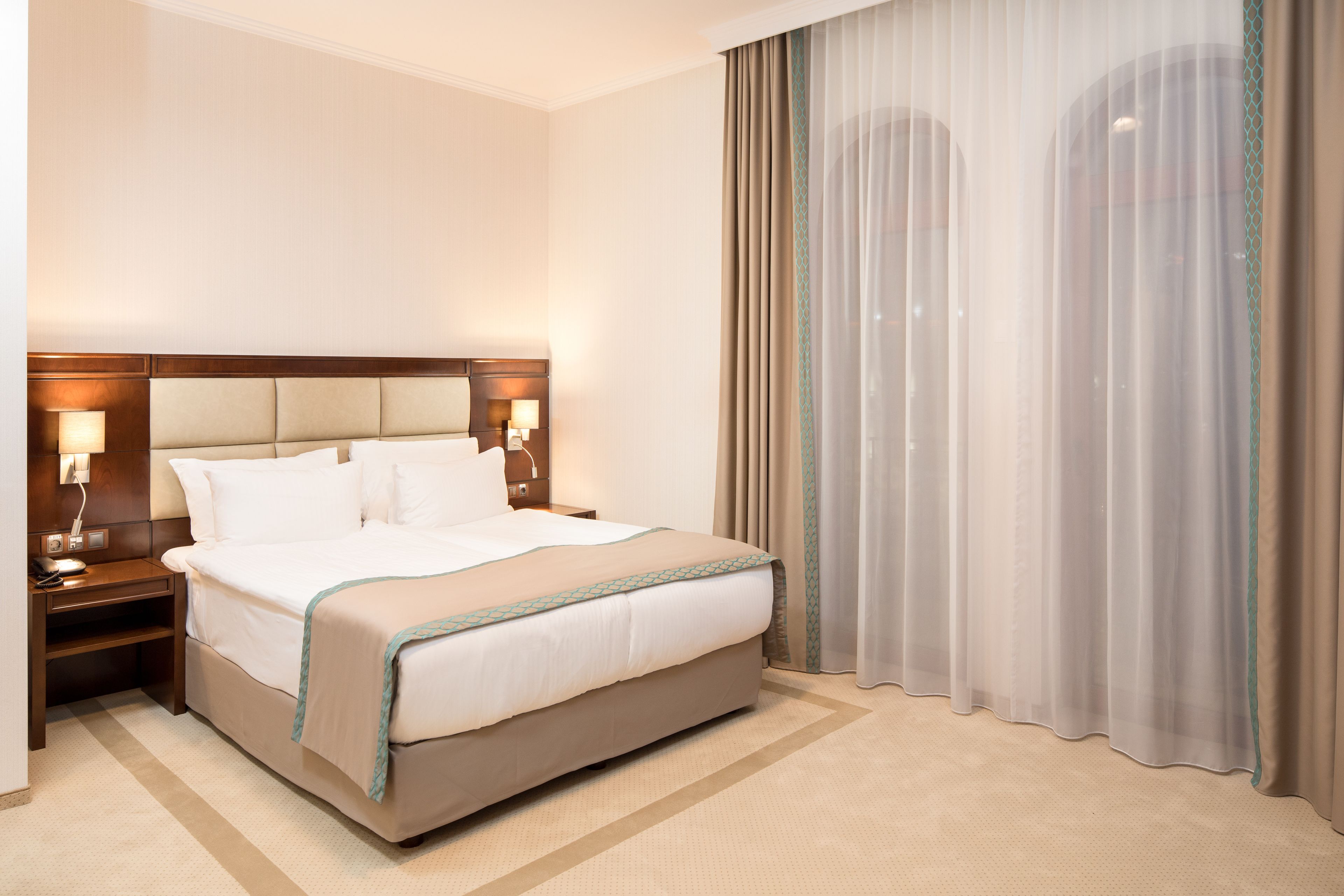 A hotel room with a large bed, bedside lamps, a nightstand, and sheer curtains.