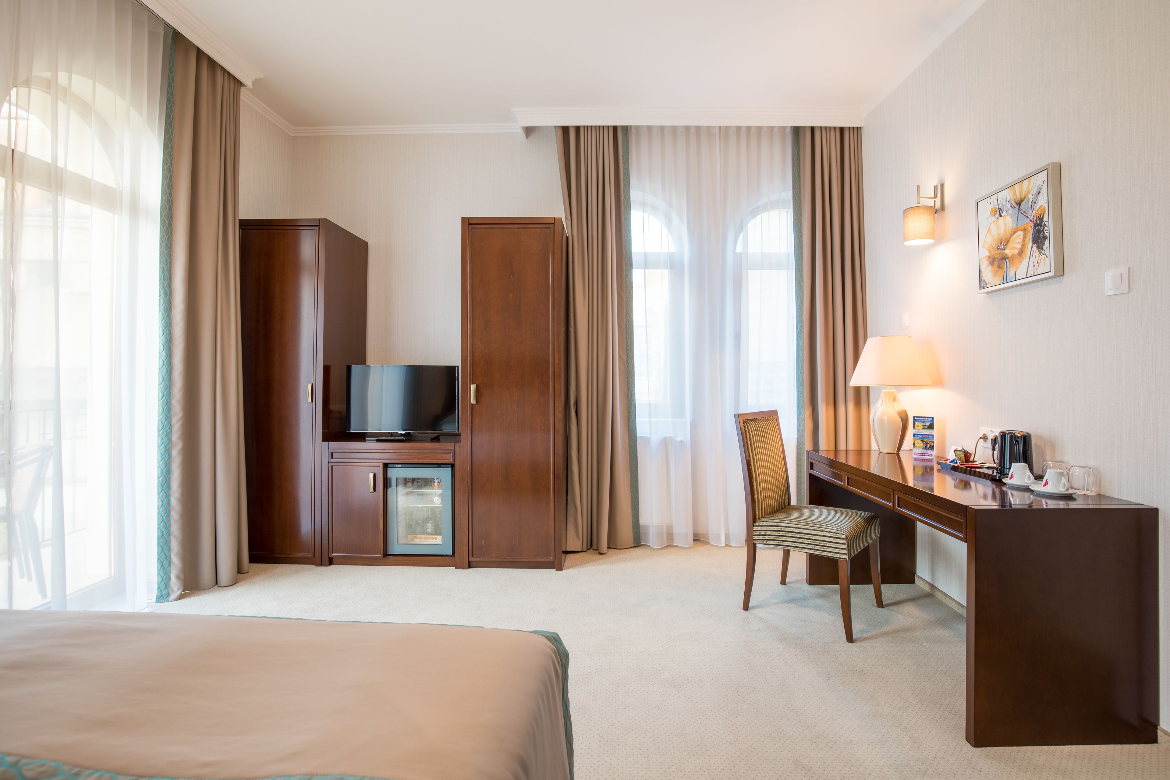 Habitación de hotel con cama, escritorio con silla, televisión, minibar y ventana con cortinas.