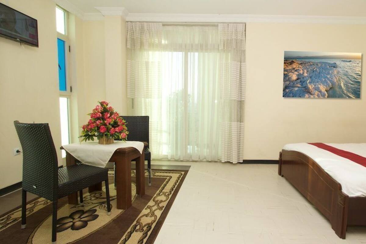 A hotel room with a bed, a desk with a chair, a window with curtains, and a painting on the wall.