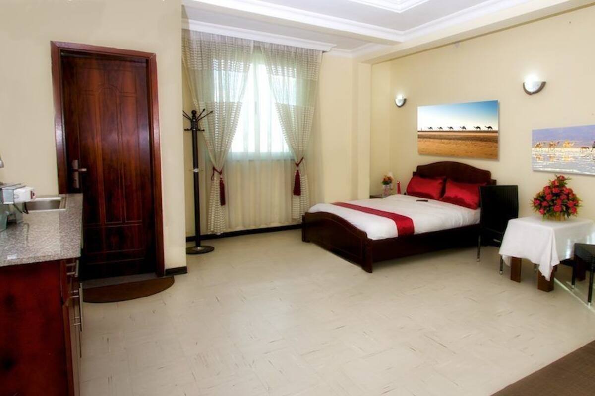 A hotel room with a bed, a desk with a chair, a small table with a vase, and a window with curtains.