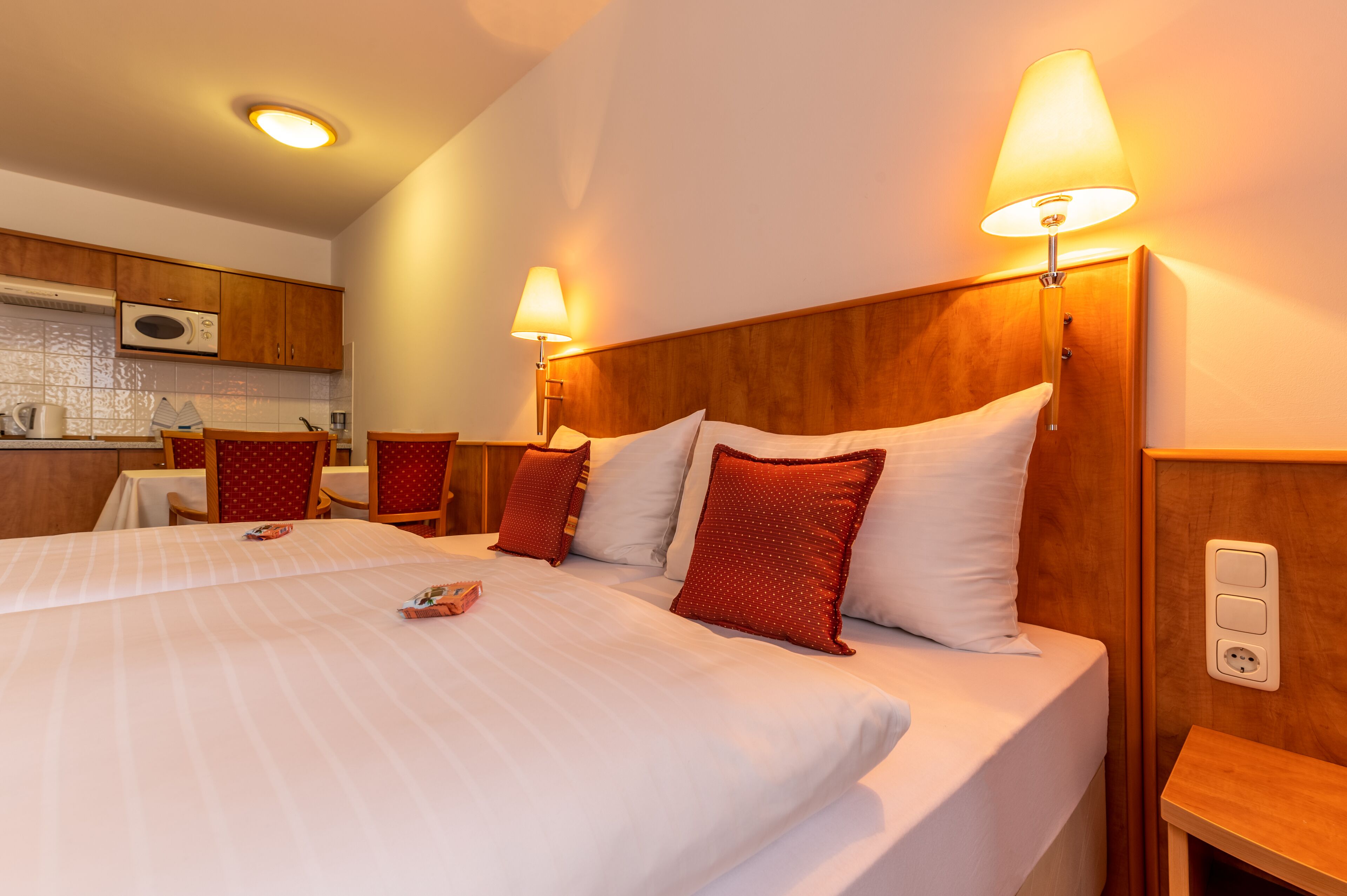 A hotel room with a large bed, two bedside lamps, a wooden headboard, a small table, and a kitchenette with a microwave.