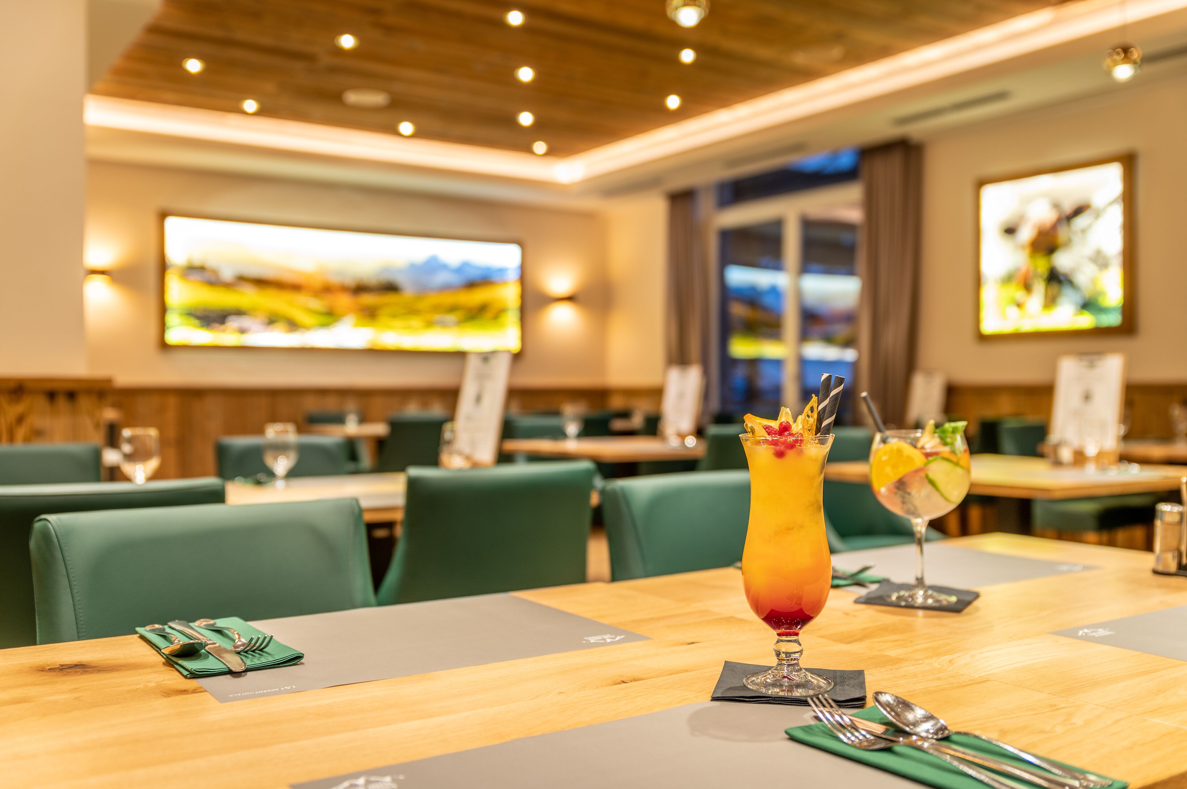 A restaurant with green upholstered chairs, wooden tables, and two cocktails on a table.