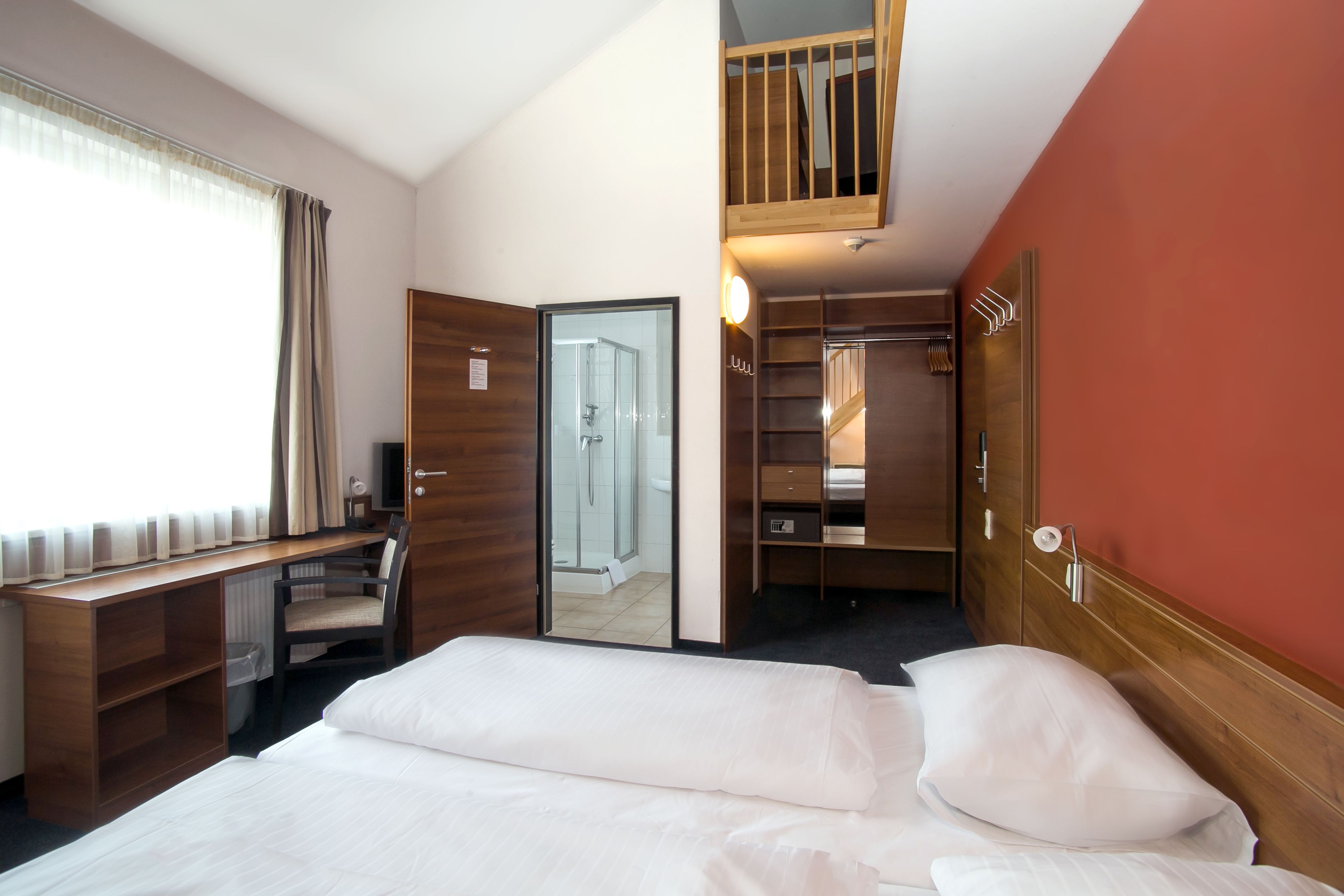 Habitación de hotel con cama, escritorio, silla, armario y baño visible a través de una puerta de cristal.