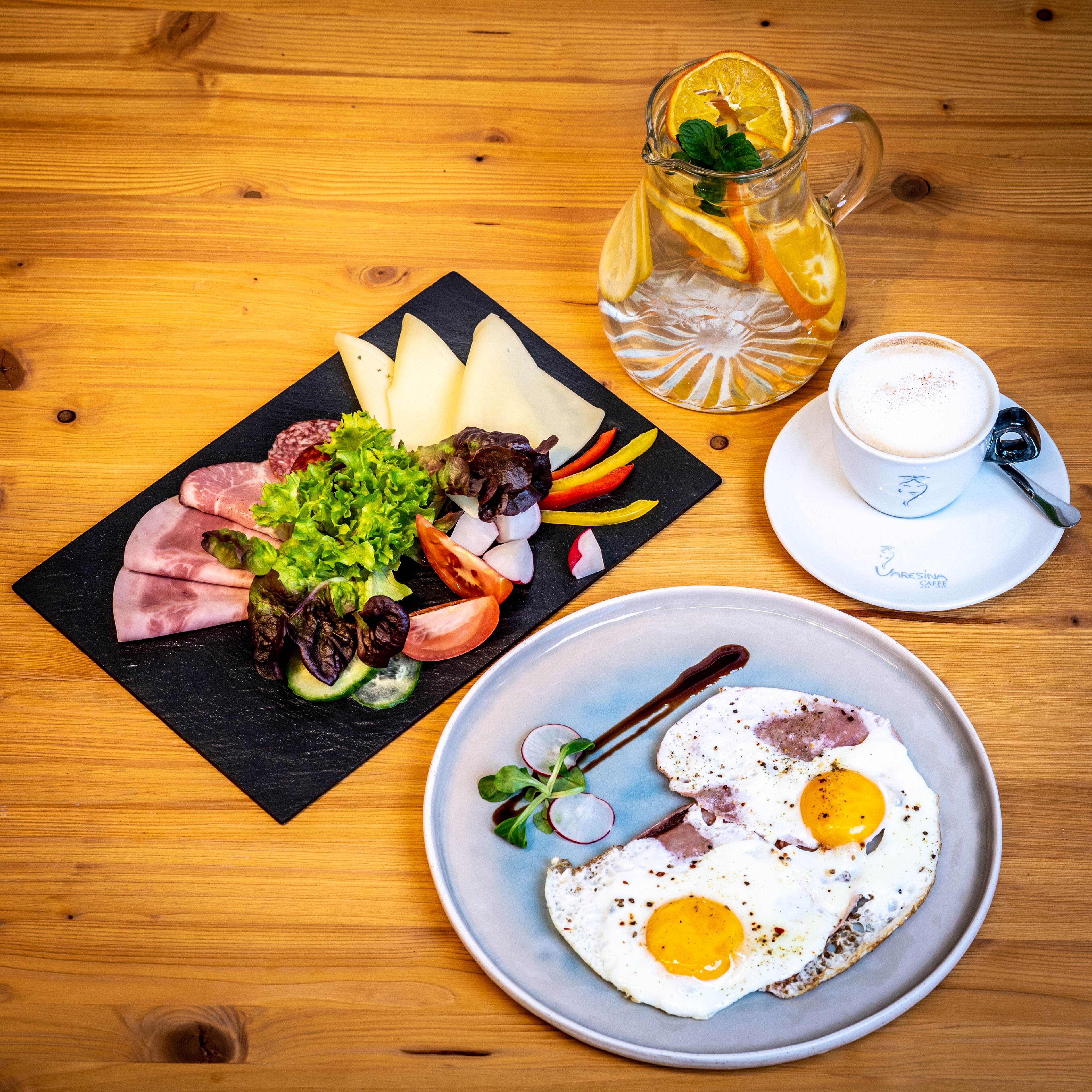 A wooden table with a meal consisting of sliced meats, vegetables, a plate with fried eggs, a glass pitcher with a slice of orange, and a cup of coffee.