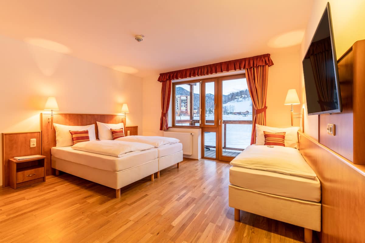 Quarto de hotel com duas camas, piso de madeira, uma janela grande com cortinas, televisão e uma mesa de cabeceira com abajur.