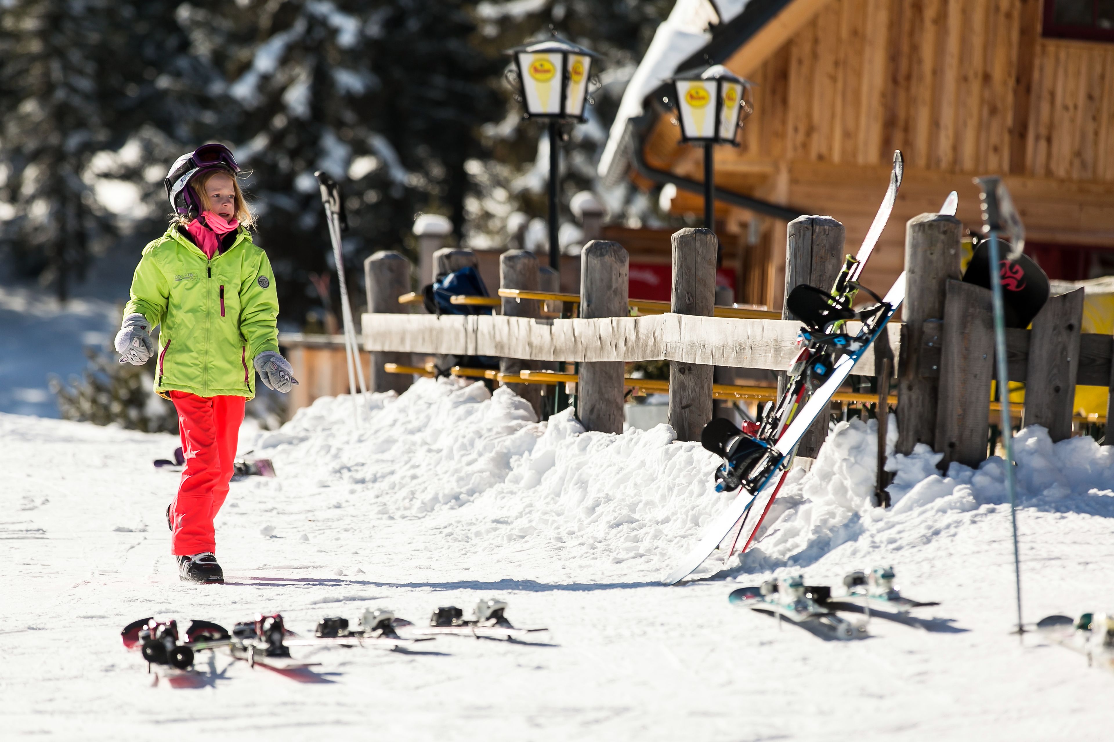 A person in a green jacket and red pants standing next to a fallen ski.