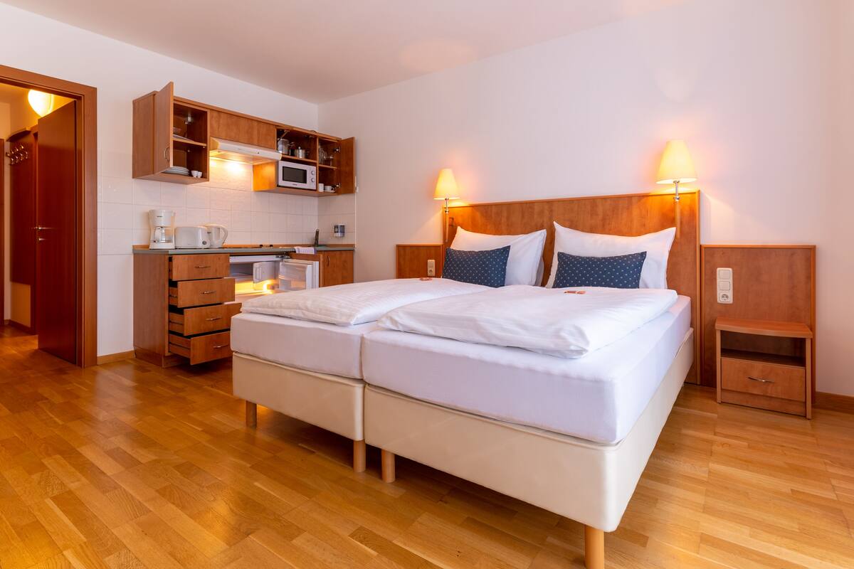 Uma cama de casal com roupa de cama branca, uma cabeceira de madeira e duas mesas de cabeceira com abajures. Cozinha compacta com micro-ondas, cafeteira e armários é visível.