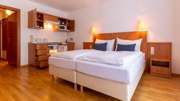 A double bed with white bedding, a wooden headboard, and two bedside tables with lamps. A kitchenette with a microwave, coffee maker, and cabinets is visible.