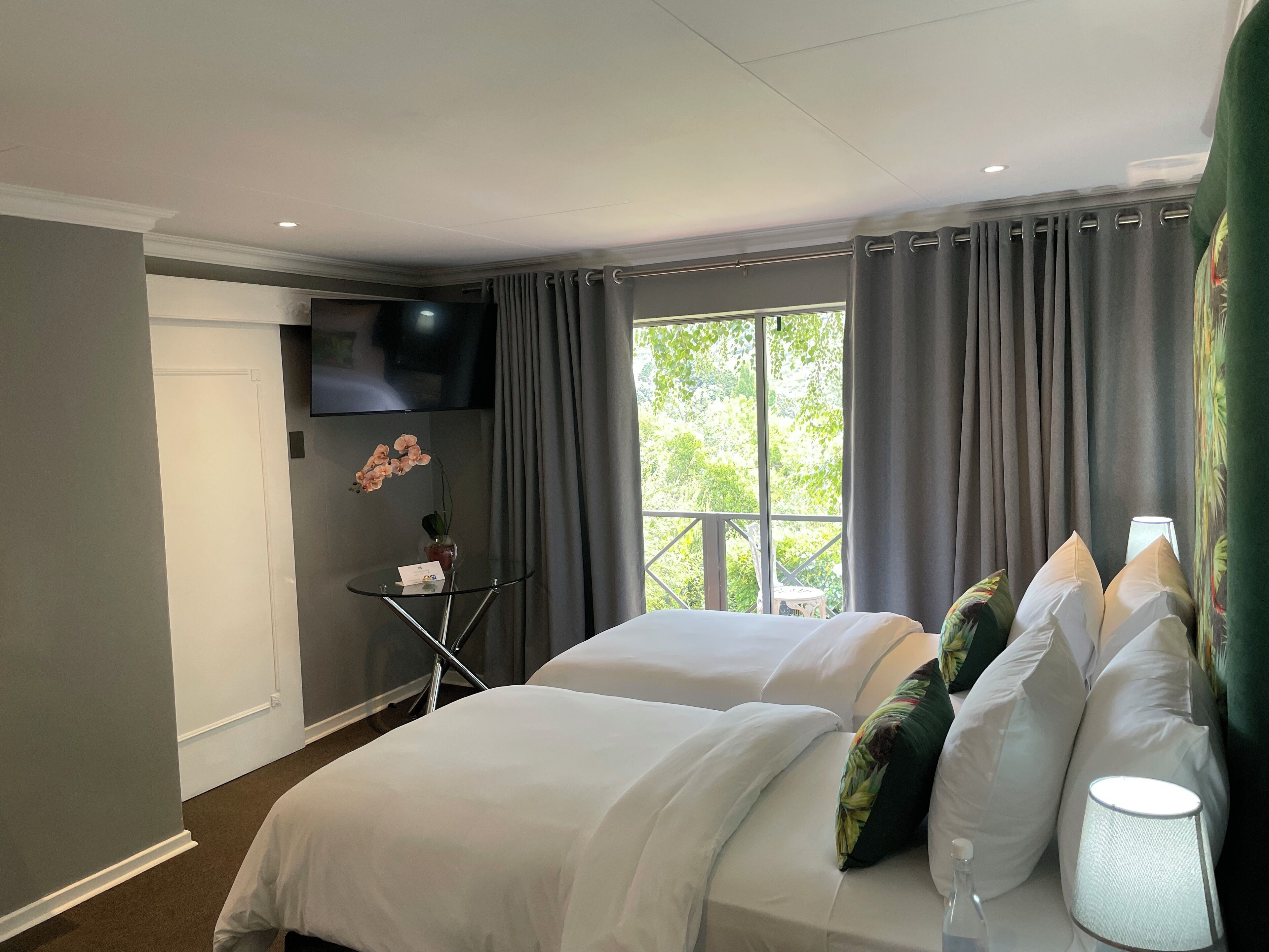 A hotel room with a large bed, a TV mounted on the wall, a small table with a vase of flowers, and a window with curtains.