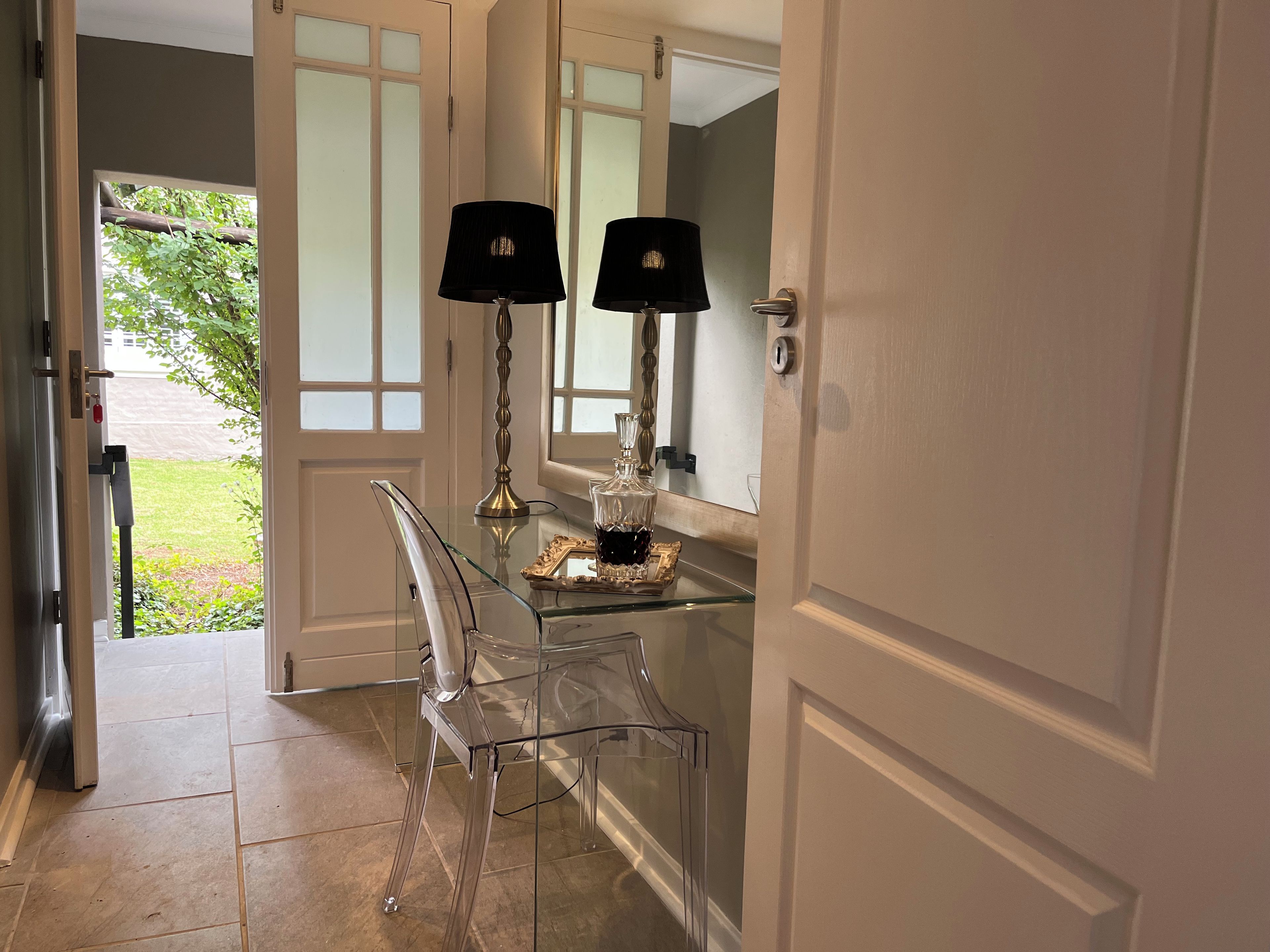 Una mesa con superficie de vidrio, dos lámparas y un objeto decorativo, una silla transparente y una puerta que da a un jardín.