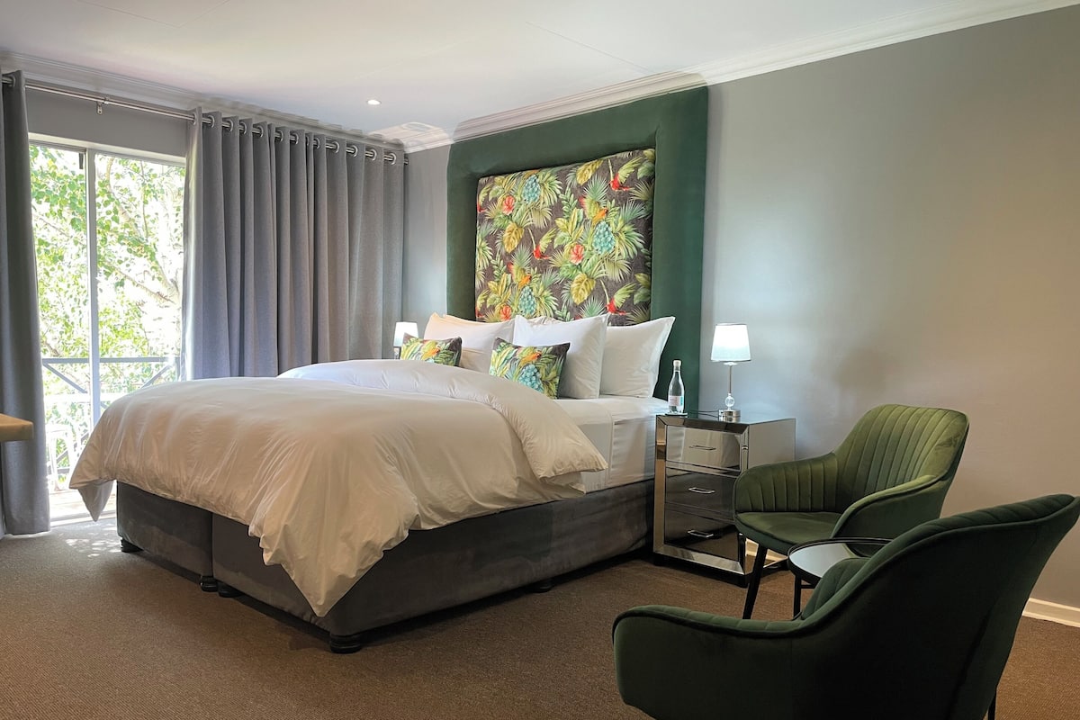 A hotel room with a large bed, a nightstand with a lamp, two green chairs, and a colorful floral headboard.