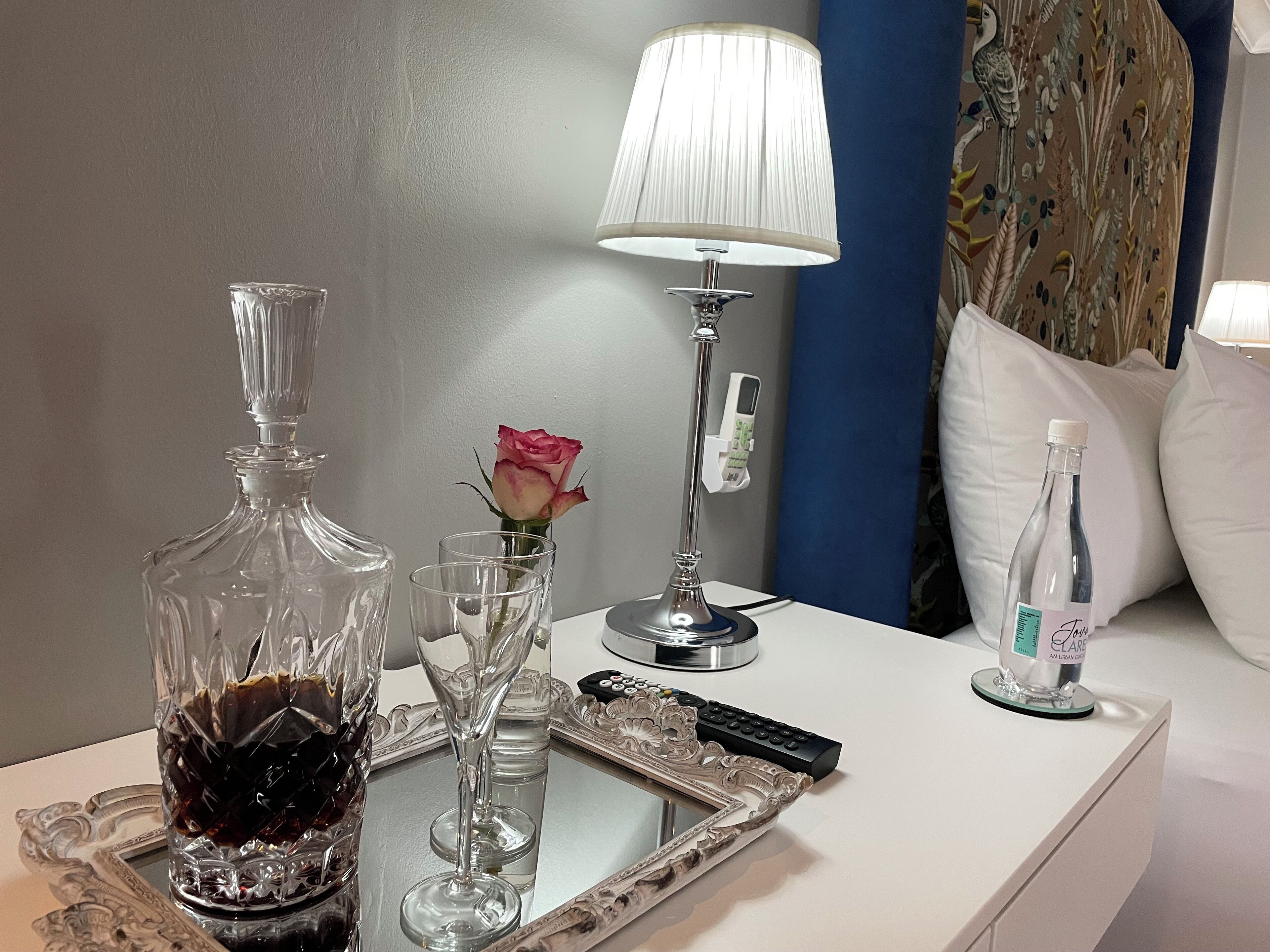 A neatly arranged table with a decanter, a rose in a glass, a remote control, and a bottle of water.