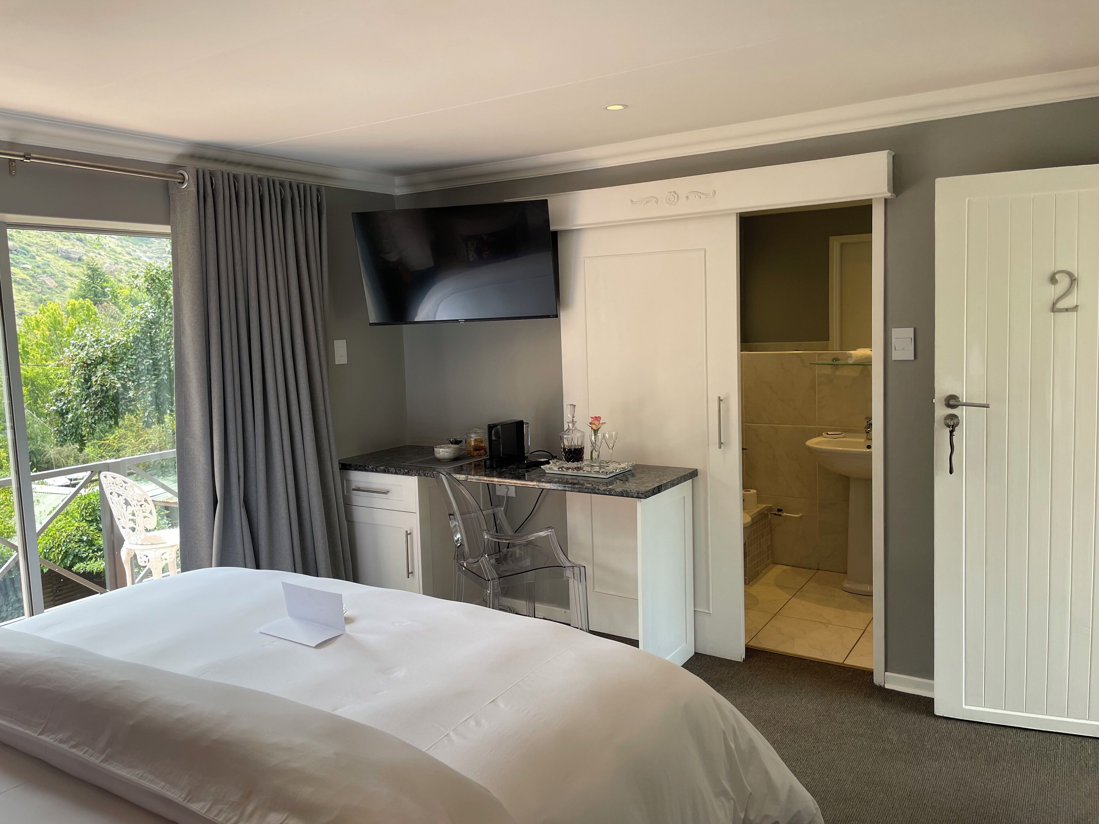 A hotel room with a bed, a desk with a TV, a bathroom, and a balcony with a chair.