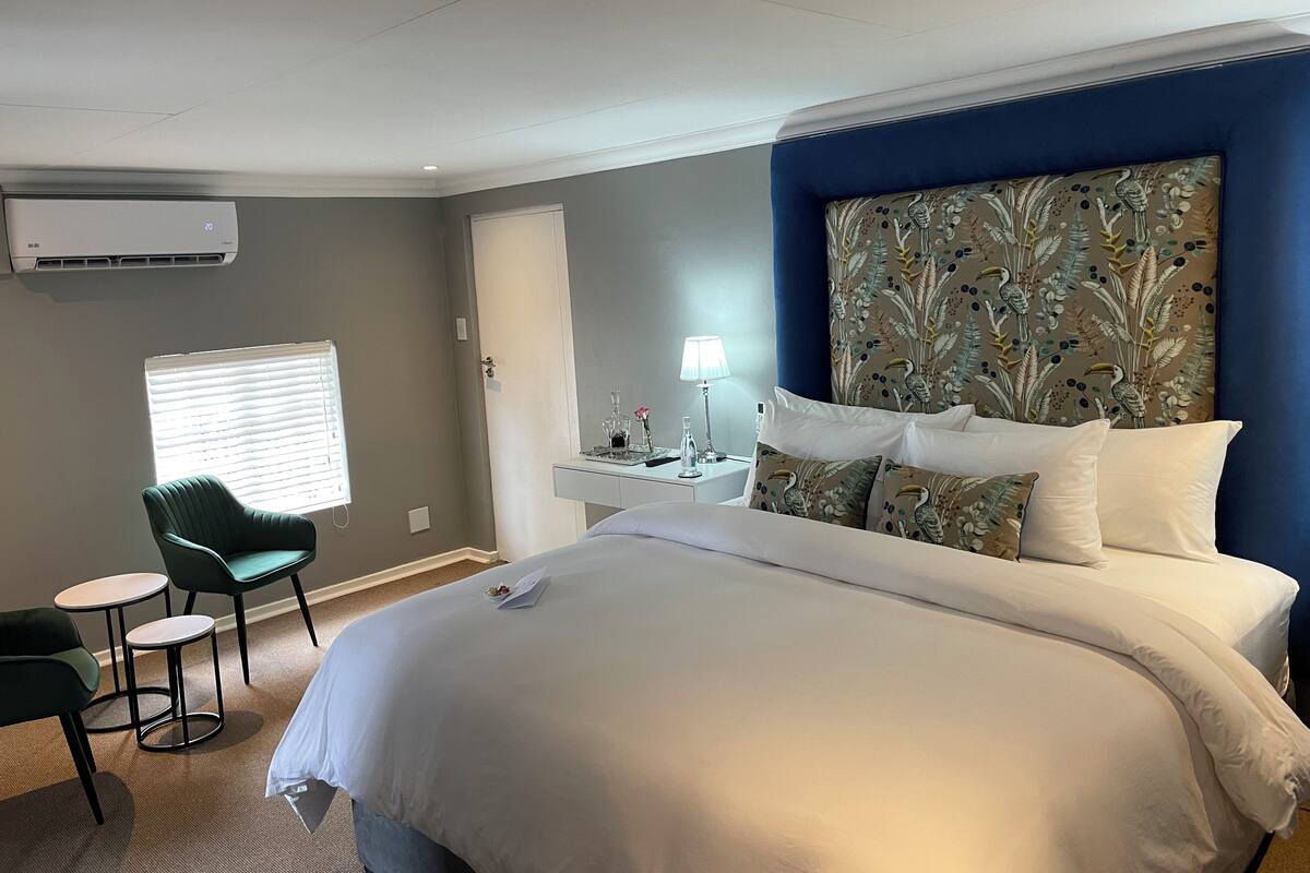 A hotel room with a large bed, a desk with a lamp, a chair, and a window with blinds.