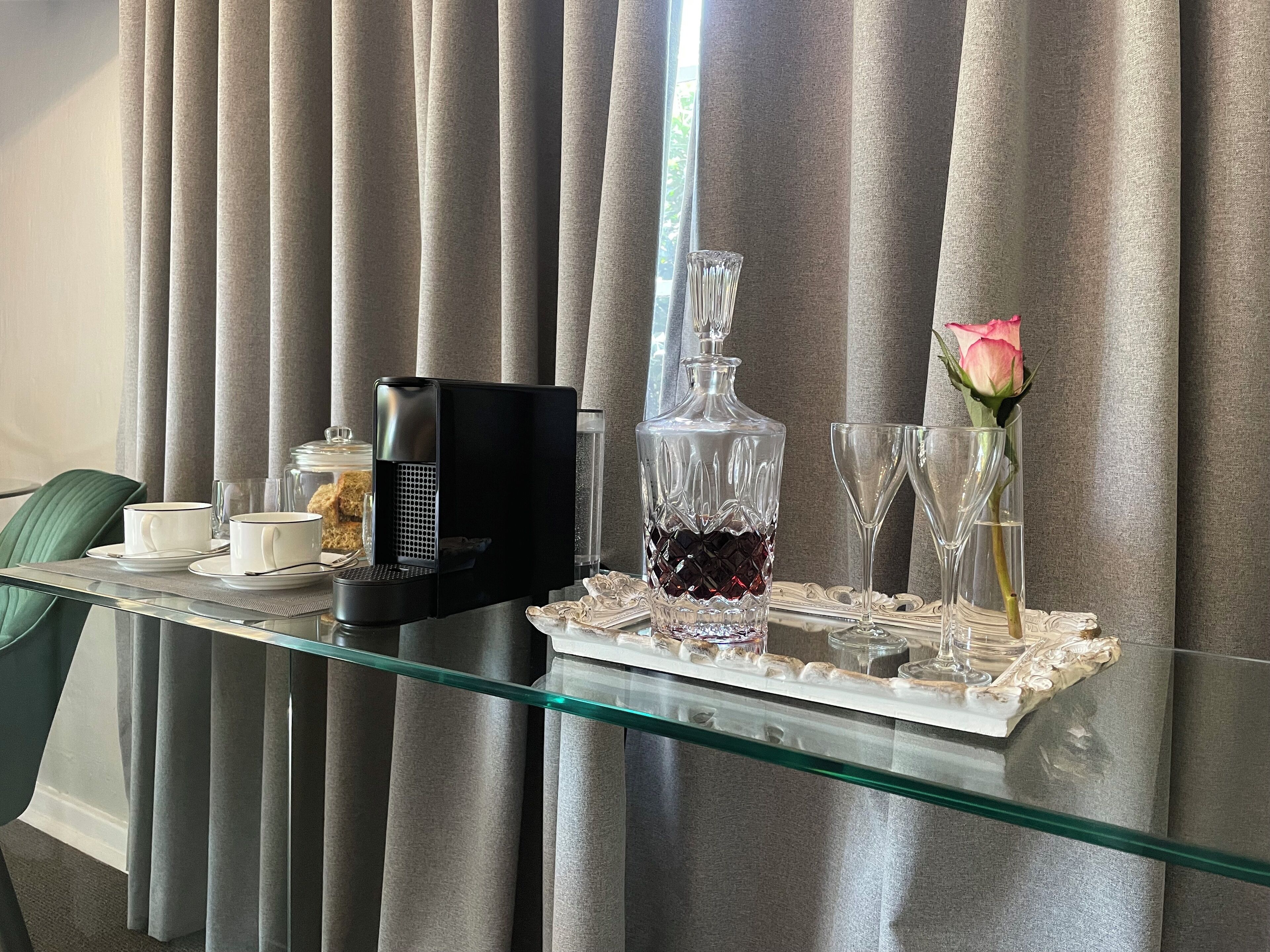 A hotel room with a glass table set for breakfast, including a coffee maker, a crystal decanter, and a vase with a rose.