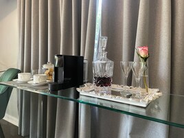 A hotel room with a glass table set for breakfast, including a coffee maker, a crystal decanter, and a vase with a rose.
