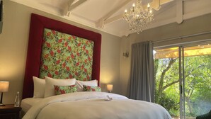 Quarto com cama grande, lustre, janela com cortinas e vista para o verde lá fora.