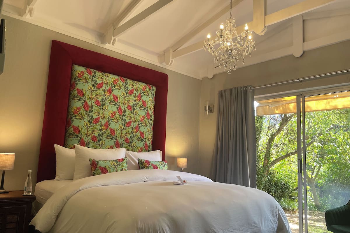 A bedroom with a large bed, a chandelier, a window with curtains, and a view of greenery outside.