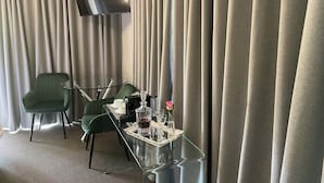 Quarto de hotel com mesa de centro de vidro, duas cadeiras verdes, televisão de tela plana na parede e uma grande janela com cortinas.