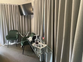 A hotel room with a glass coffee table, two green chairs, a flat-screen TV mounted on the wall, and a large window with curtains.