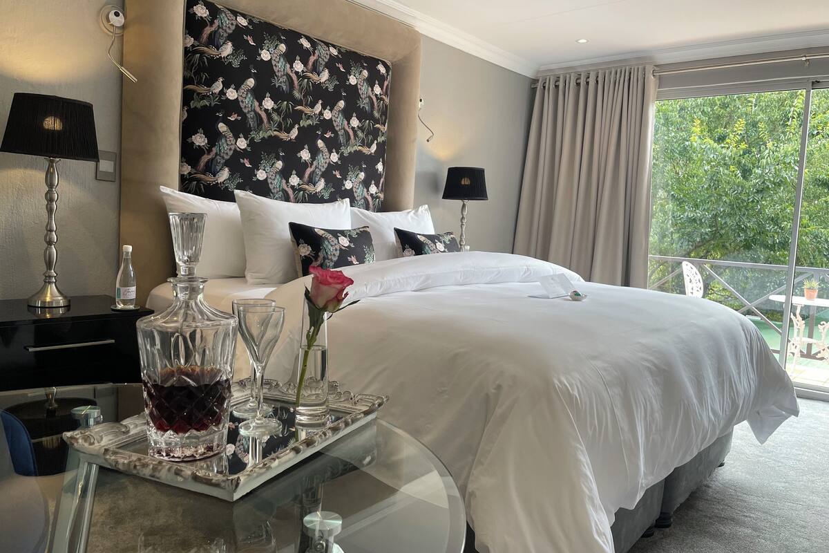 A hotel room with a large bed, a glass table with a tray, a vase with a rose, and a view of greenery outside.
