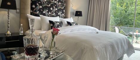 A hotel room with a large bed, a glass table with a tray, a vase with a rose, and a view of greenery outside.