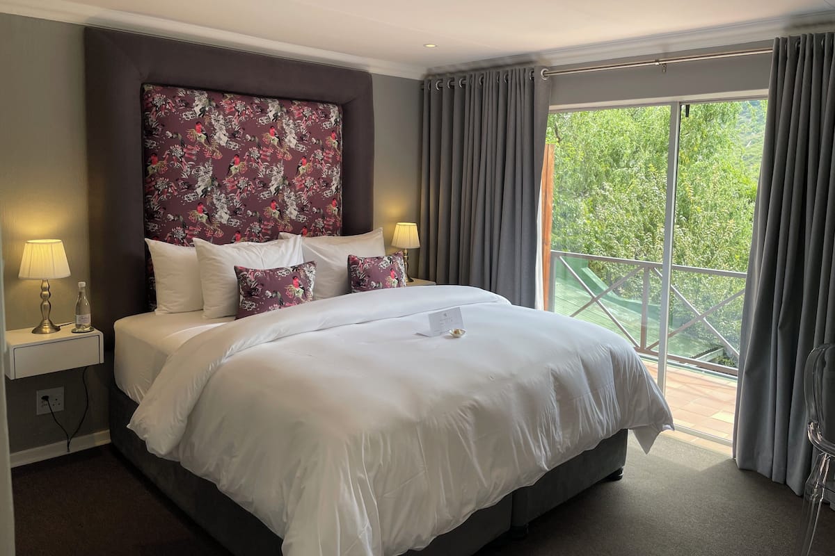 A hotel room with a large bed, two bedside lamps, a floral headboard, and a balcony with a view of greenery.