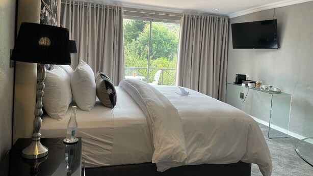 Quarto de hotel moderno com uma cama grande, televisão de tela plana, mesa de cabeceira com abajur e vista para o verde pela janela.