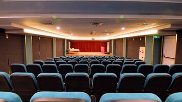 Sala de conferências com fileiras de cadeiras voltadas para um palco com cortina vermelha.