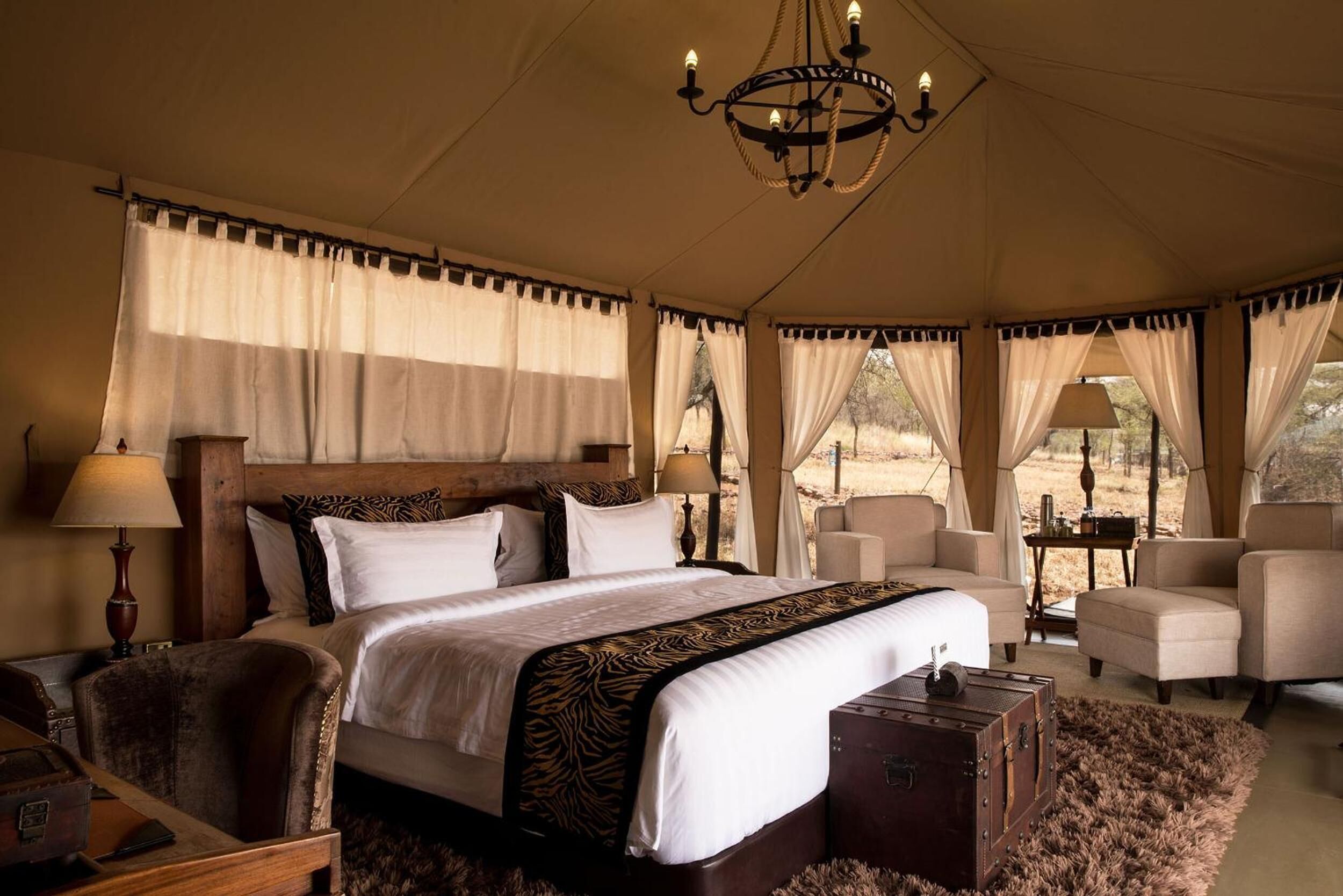 A tented room with a large bed, two chairs, a wooden nightstand, and a chandelier.