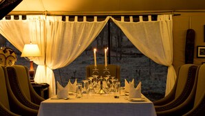 A dining table set for two with candles, a lamp, and a view of a snowy landscape through sheer curtains. - Ole Serai Luxury Camp (Bariadi)
