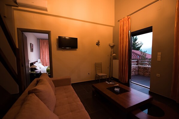 A living room with a sofa, a coffee table, a TV mounted on the wall, a chair, and a view of the outside through a door. - Melies Hotel (Sparta)