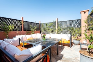 Un patio al aire libre con una mesa de vidrio, asientos blancos acolchados y plantas en macetas.