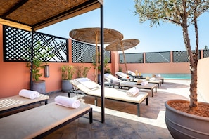 Un espacio lounge al aire libre iluminado por el sol, con sillones, sombrillas y plantas en macetas.
