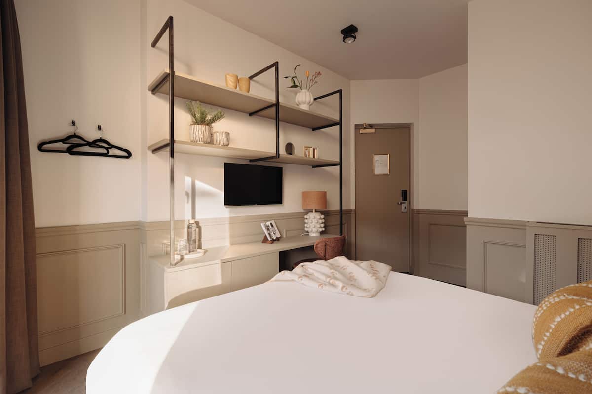 A hotel room with a bed, a TV mounted on the wall, a desk with a lamp, and shelves with decorative items.