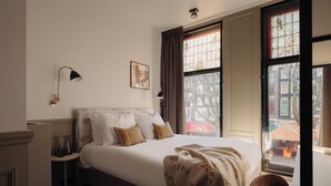 A hotel room with a large bed, a bedside table, a lamp, and a window with a view of the city. - Adam Downtown (Amsterdam)