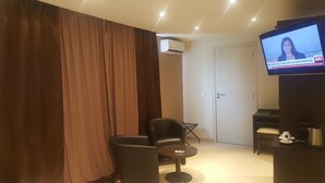 A hotel room with a television displaying a news channel, a seating area with a chair and a small table, and a door in the background. - Hôtel M'lys (Conakry)