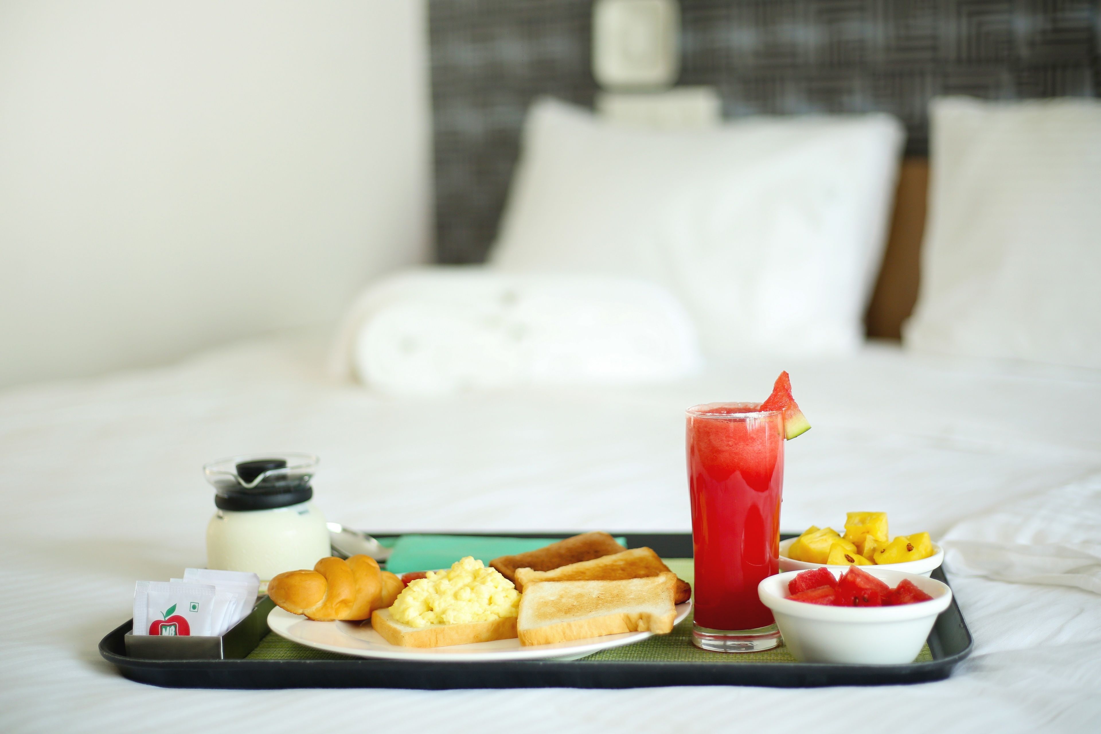 A tray with breakfast items including toast, scrambled eggs, a glass of red juice, and fruit on a bed.