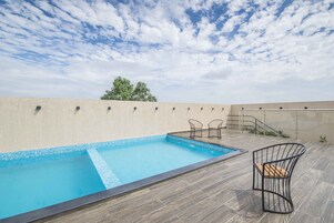 Una piscina en la azotea con borde de baldosas, rodeada por una terraza de madera y dos sillas de alambre negro.